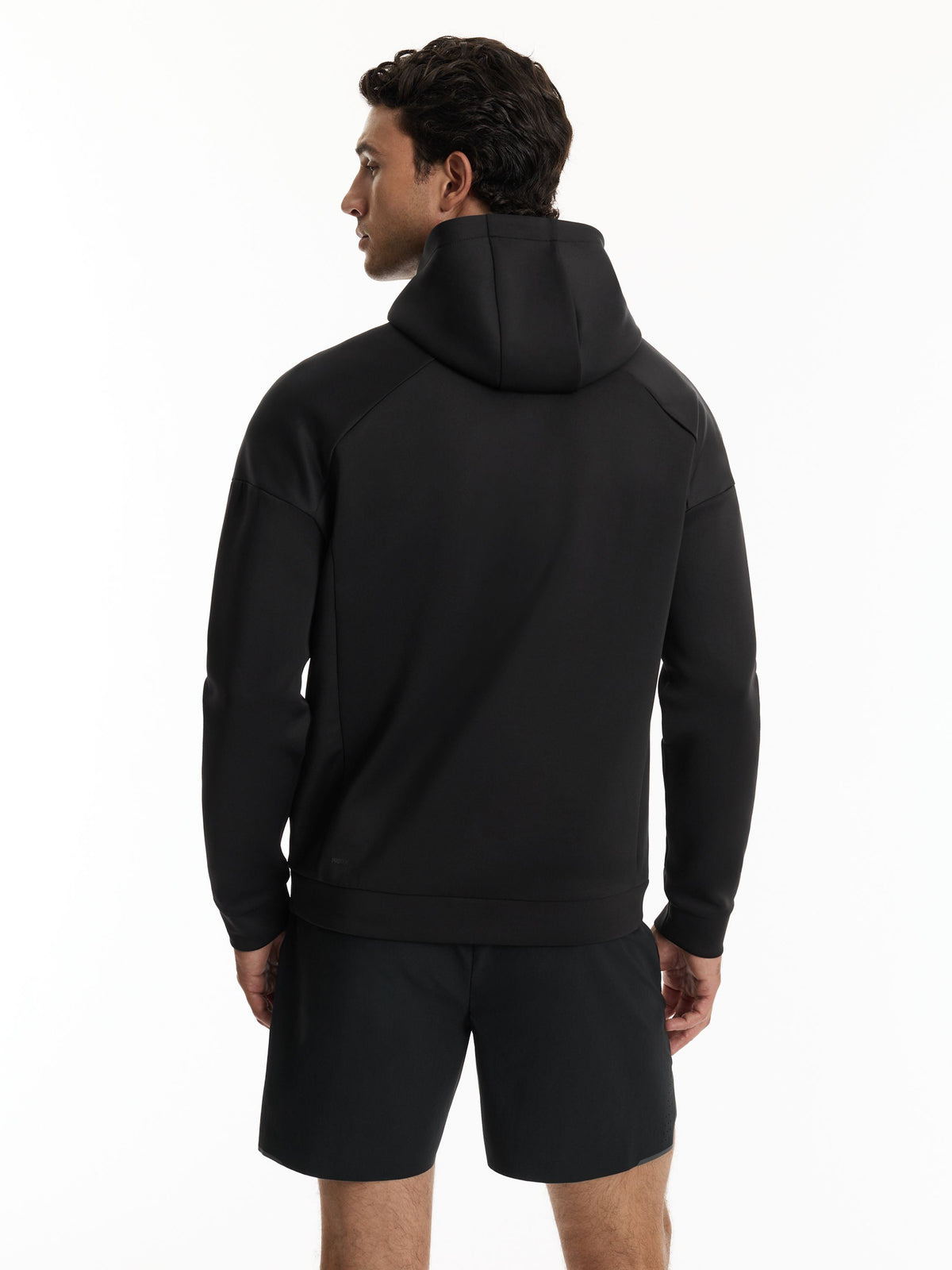 A man wearing a black hooded sweatshirt and matching black shorts, shown from the back against a plain white studio background.