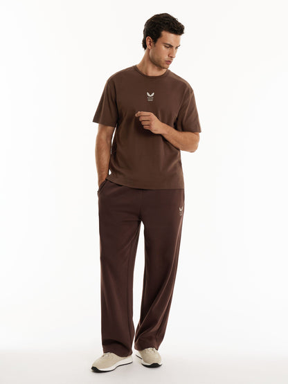 A man modeling a matching brown short-sleeve t-shirt and sweatpants set with a small white Adidas logo on the chest and thigh, photographed against a clean white studio background.