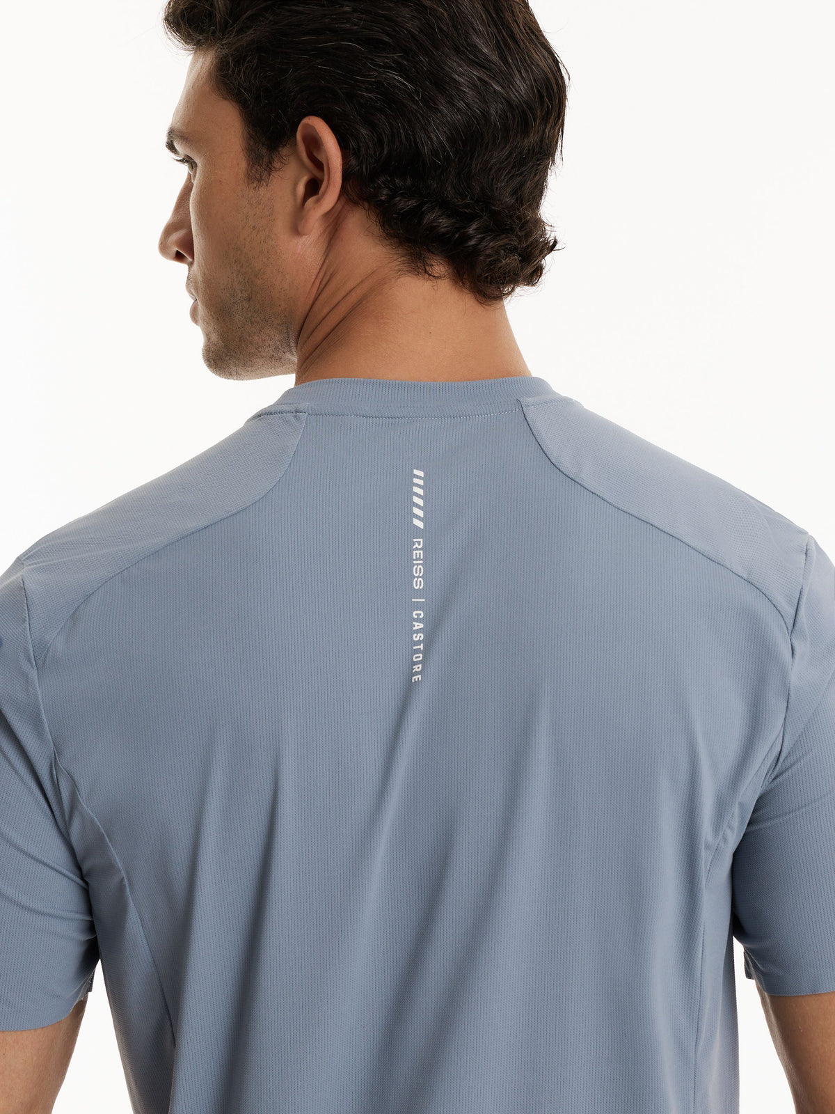 Rear view of a man wearing a light blue-gray athletic t-shirt with a vertical white 'REISS | CASTORE' logo printed on the back.