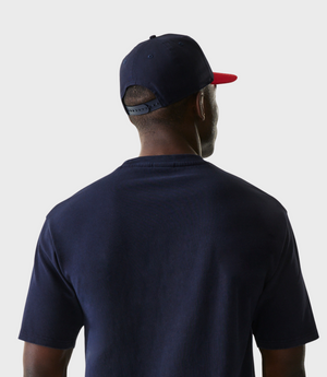Rear three quarter view of a man wearing a navy cap with a red brim and a navy Oracle Red Bull Racing T shirt.