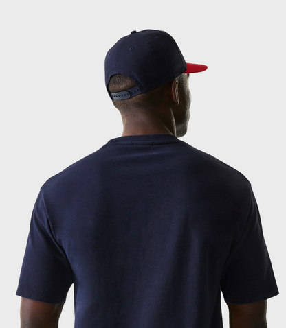 Rear three quarter view of a man wearing a navy cap with a red brim and a navy Oracle Red Bull Racing T shirt.
