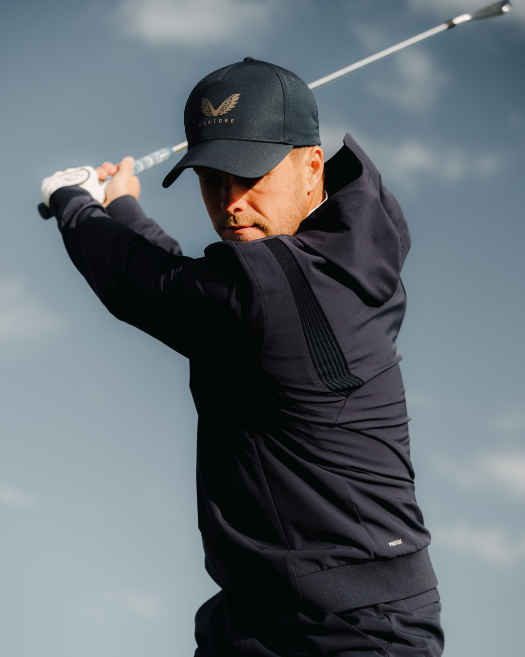 Golfer wearing the Golf Pinnacle Performance Hoodie in midnight navy mid swing with a club
