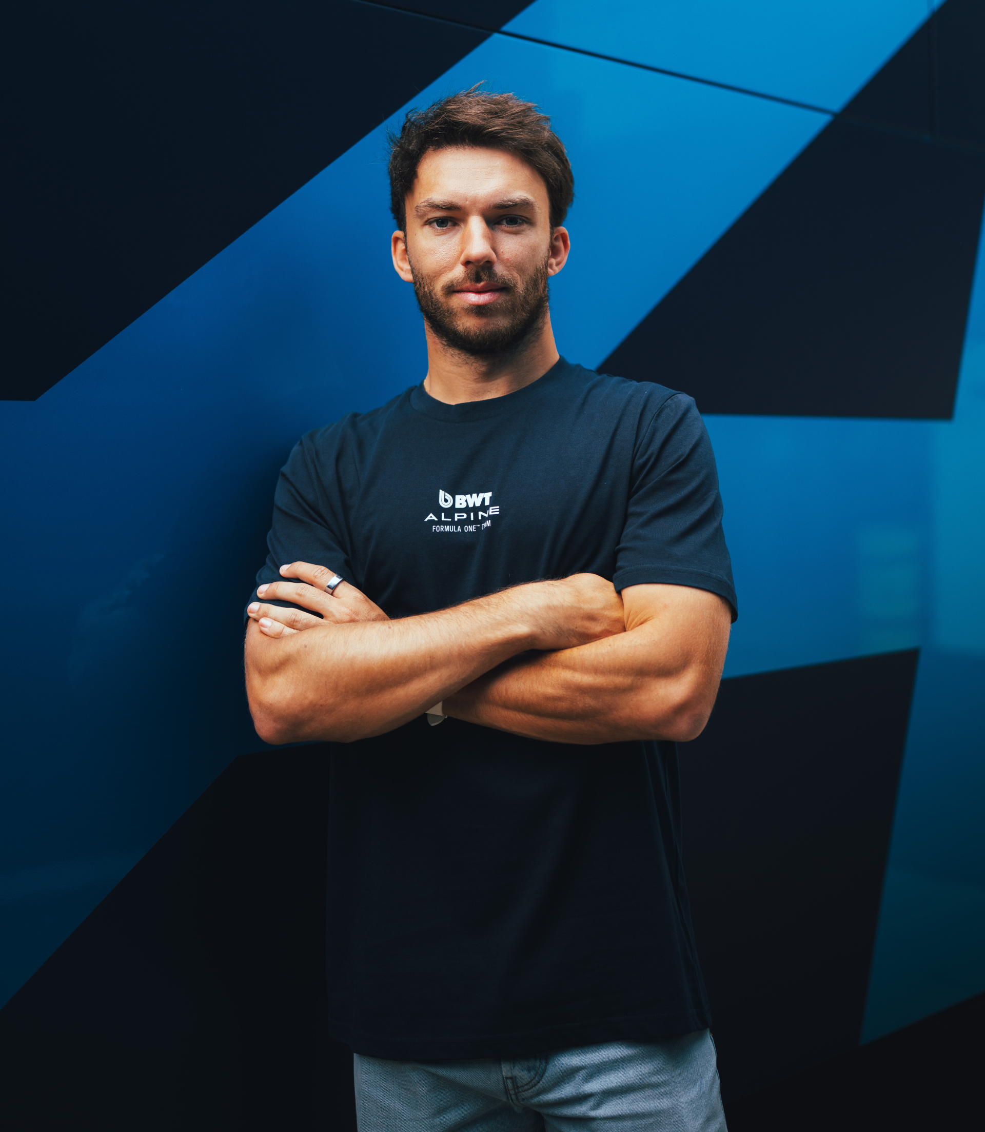 Portrait of a man in a navy BWT Alpine T shirt with logo on chest and arms crossed against a blue geometric wall.