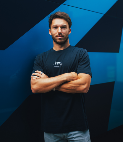 Portrait of a man in a navy BWT Alpine T shirt with logo on chest and arms crossed against a blue geometric wall.