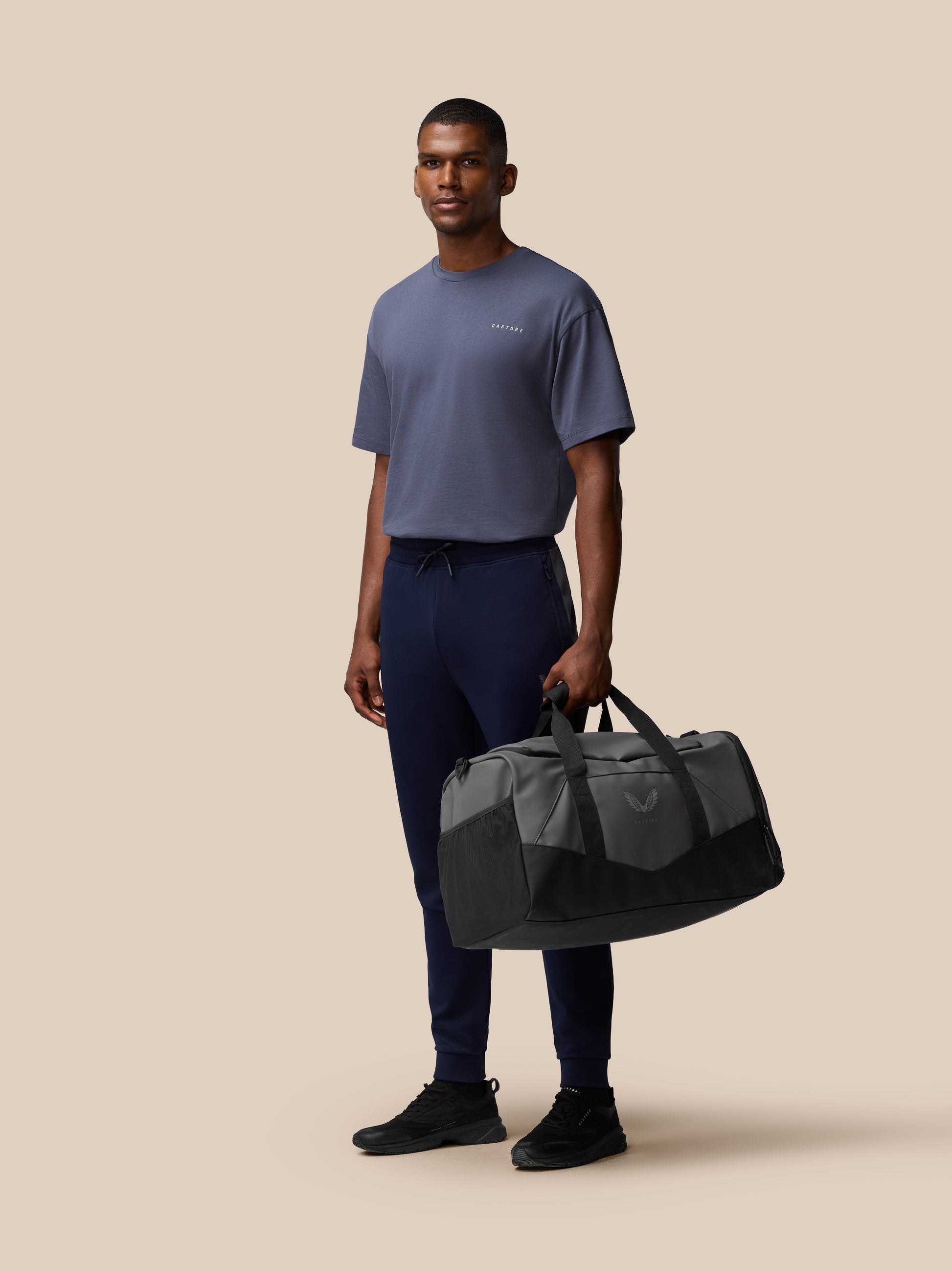 Male model wearing a blue gray tee stands with a gunmetal and black Core Holdall by his side