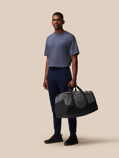 Male model wearing a blue gray tee stands with a gunmetal and black Core Holdall by his side