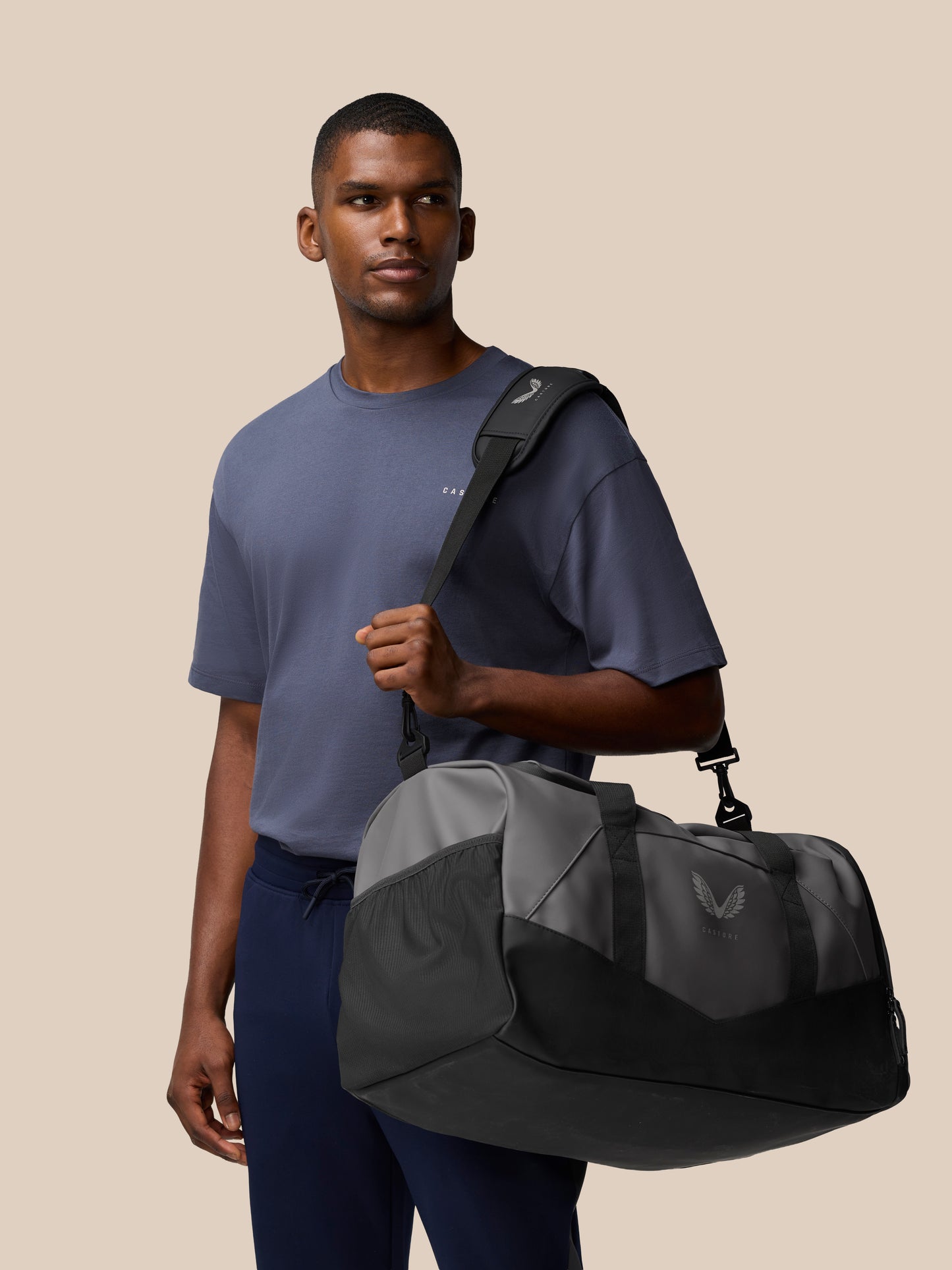 Male model in a blue tee carries a gunmetal and black Core Holdall over his shoulder with the strap across his chest