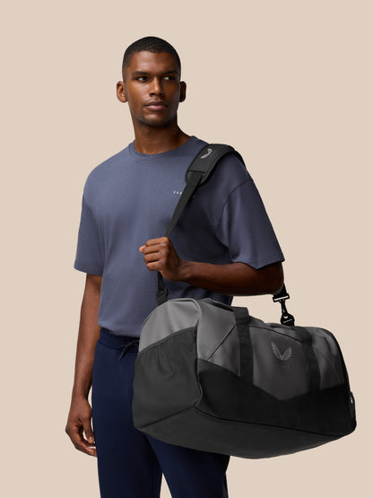 Male model in a blue tee carries a gunmetal and black Core Holdall over his shoulder with the strap across his chest