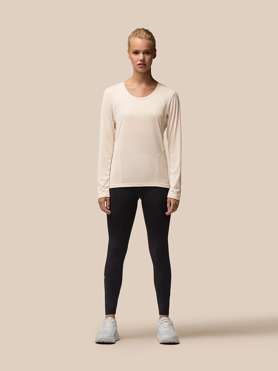 Front view of a model in beige Flow Movement long sleeve top paired with black leggings and white sneakers.