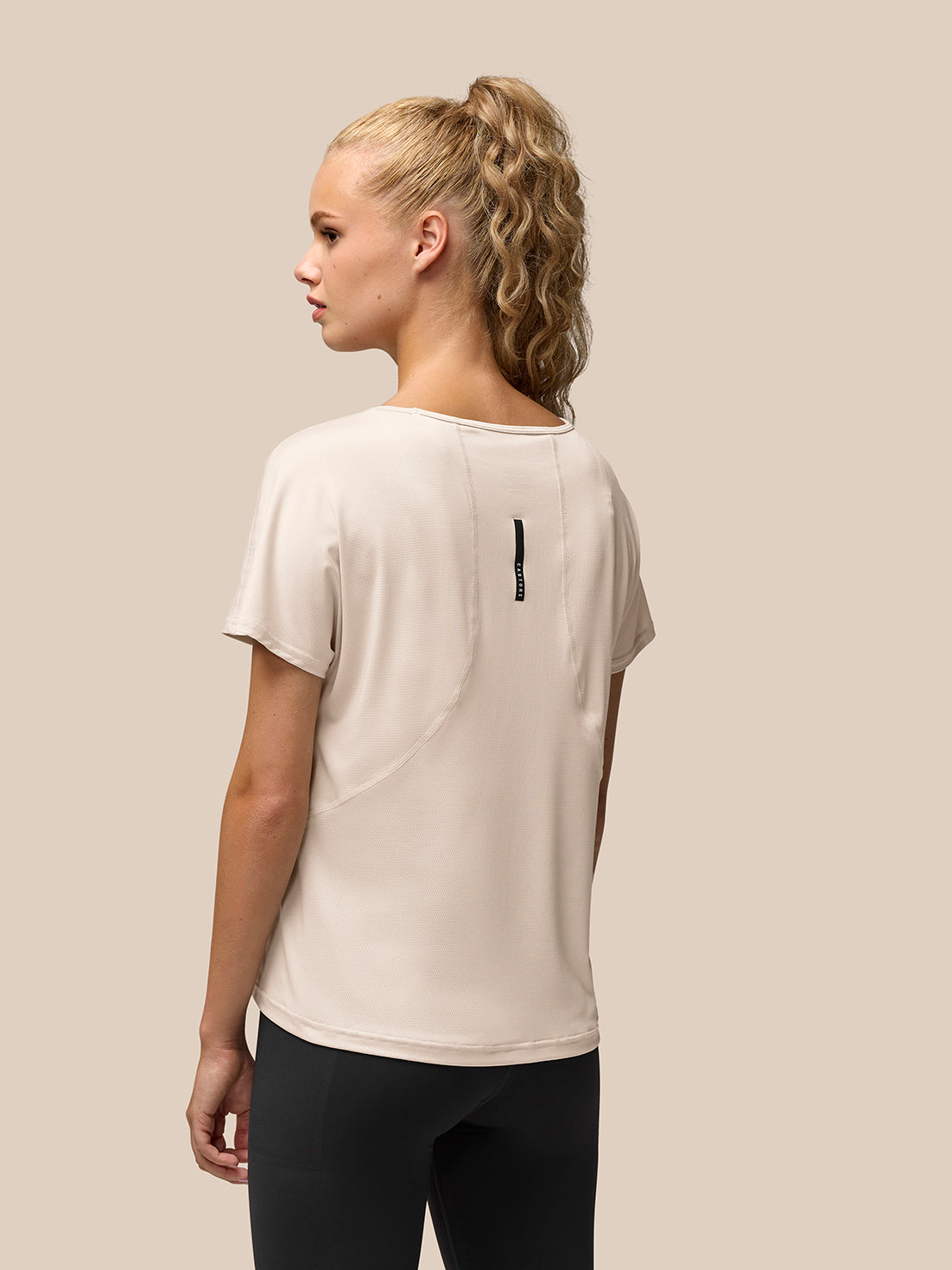 Back view of a beige Flow Training T-Shirt on a model, featuring a vertical black logo stripe running down the upper back.
