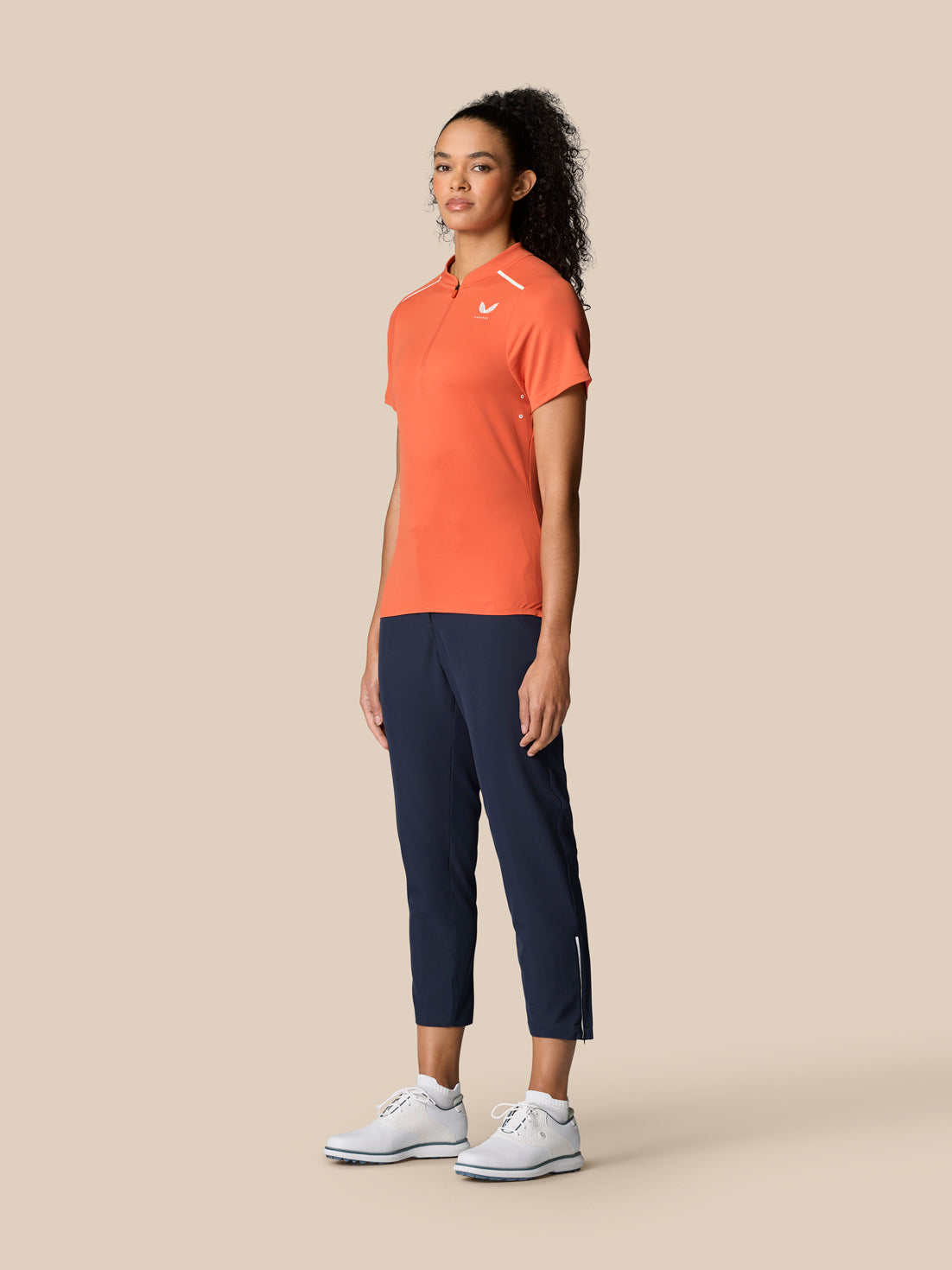 Full-length view of a female model wearing a Deep Coral Pinnacle Varsity Polo with navy pants and white sneakers