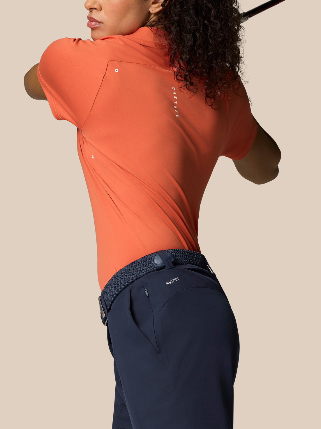 Back view of a woman wearing a Deep Coral Pinnacle polo with Castore branding, navy pants, and woven belt.