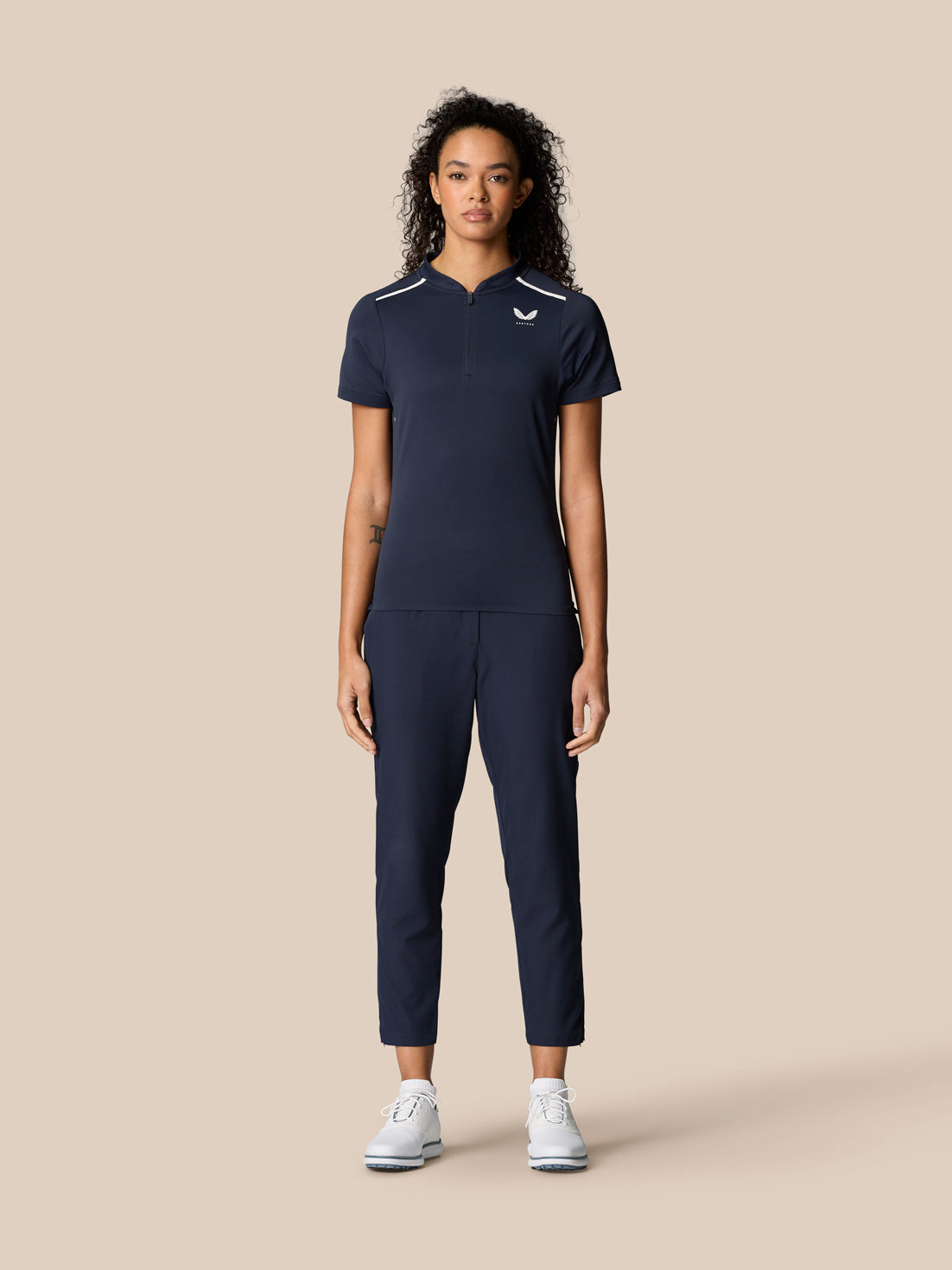 Woman wearing navy Pinnacle Polo with front quarter zip and white piping, paired with navy pants and white sneakers.