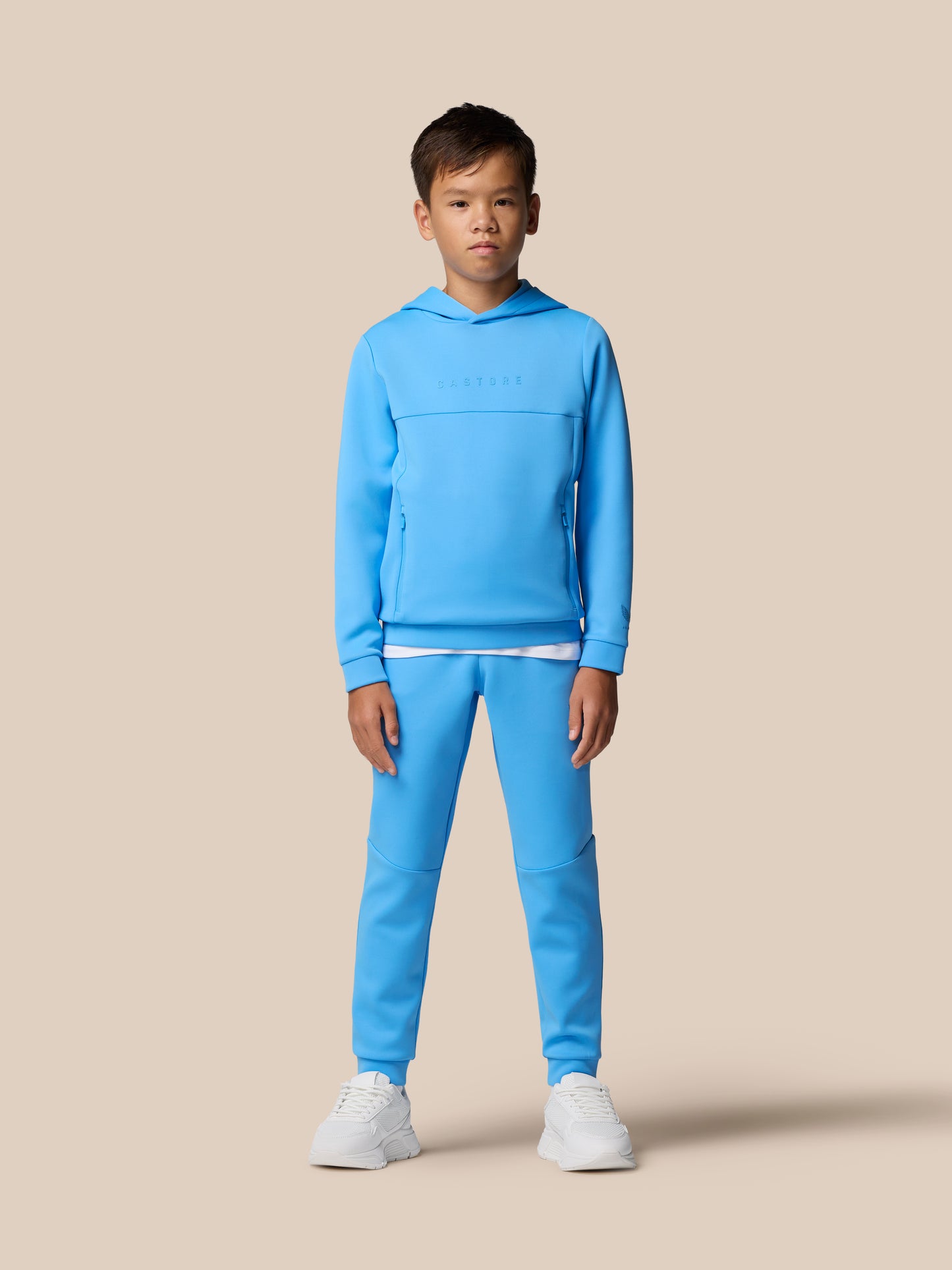 Young model wearing a blue junior Flex Scuba tracksuit with hooded top and matching joggers against a beige backdrop