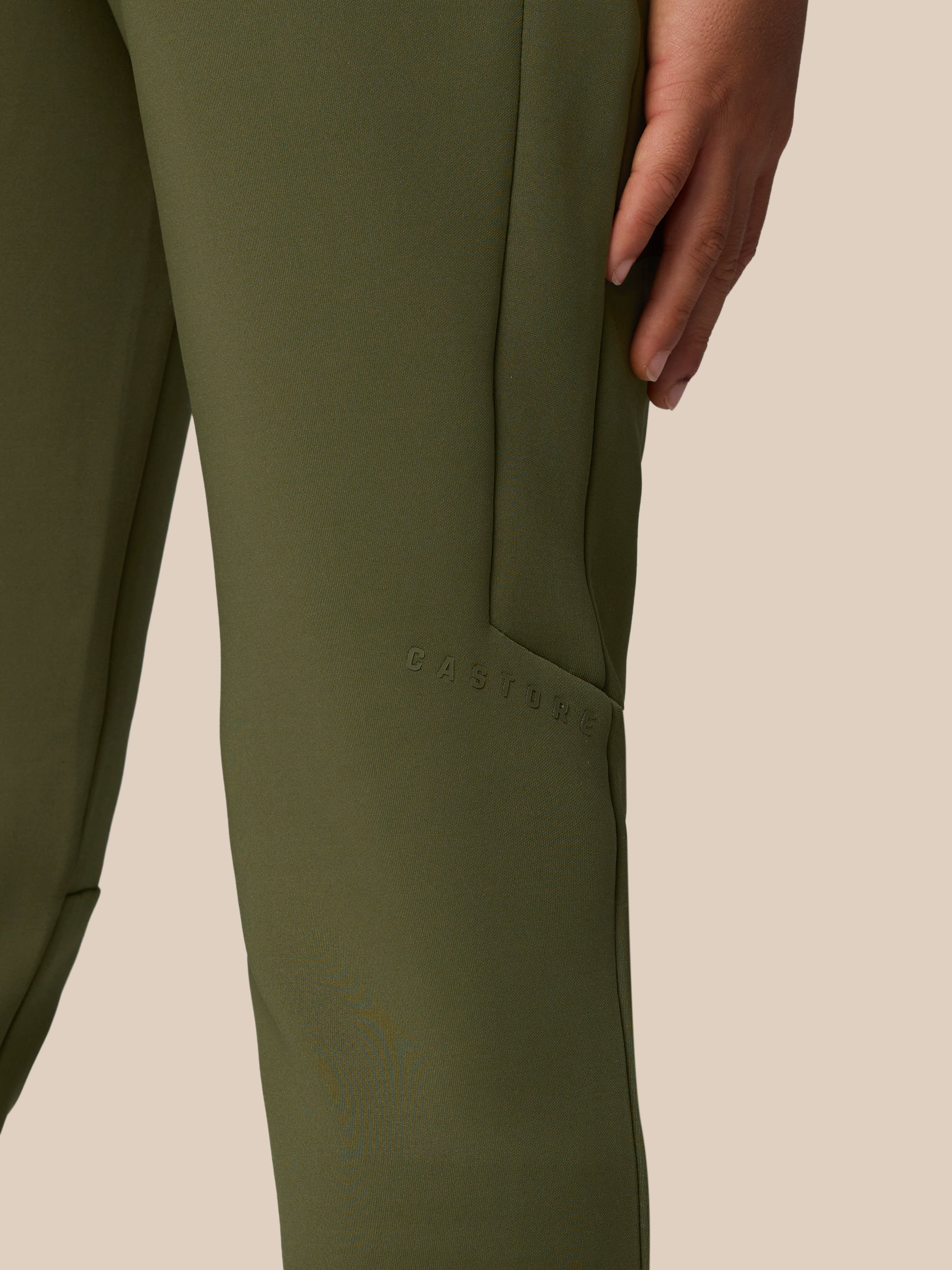 Close-up of olive green scuba fabric on a leg from Junior’s Flex Scuba Trousers, with a hand at the side and Castore branding embossed along the seam.