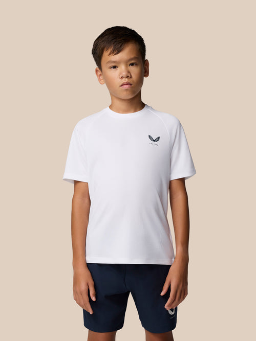 Young boy wearing a white Junior's Adapt Training T-Shirt with Castore logo on the chest and navy shorts against a beige backdrop