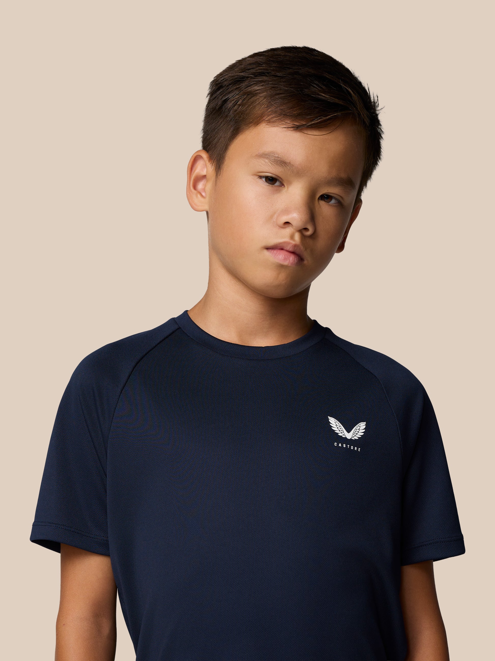 Portrait of a young boy in a navy Castore Adapt short sleeve shirt with a white Castore logo on the chest against a beige background.