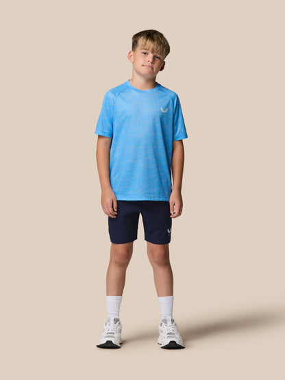 Full body shot of a boy in blue geometric print short sleeve T shirt and navy shorts with Castore logo on left chest against a beige backdrop
