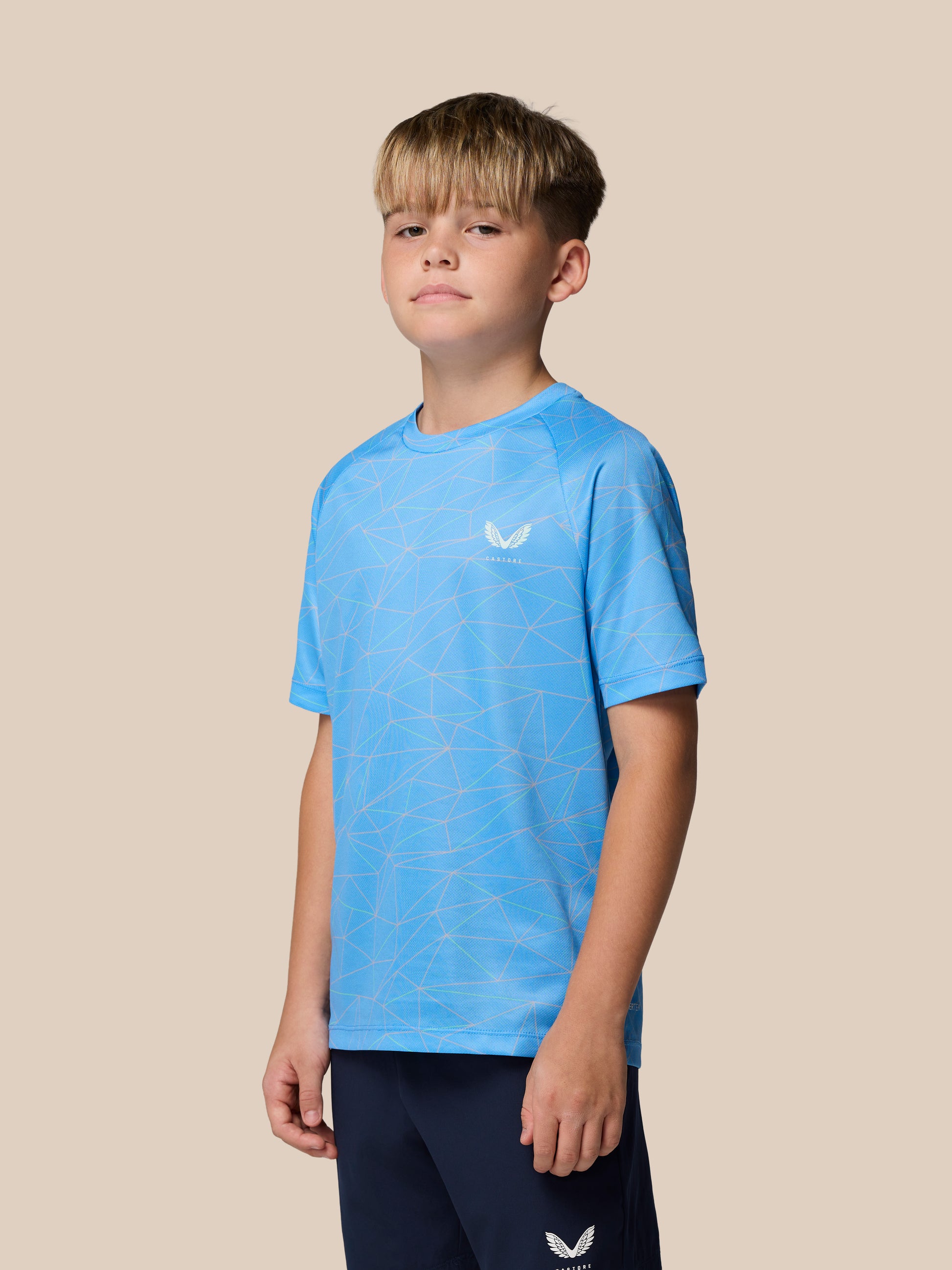 Young boy wearing light blue geometric print Castore short sleeve T shirt with left chest logo against a beige backdrop