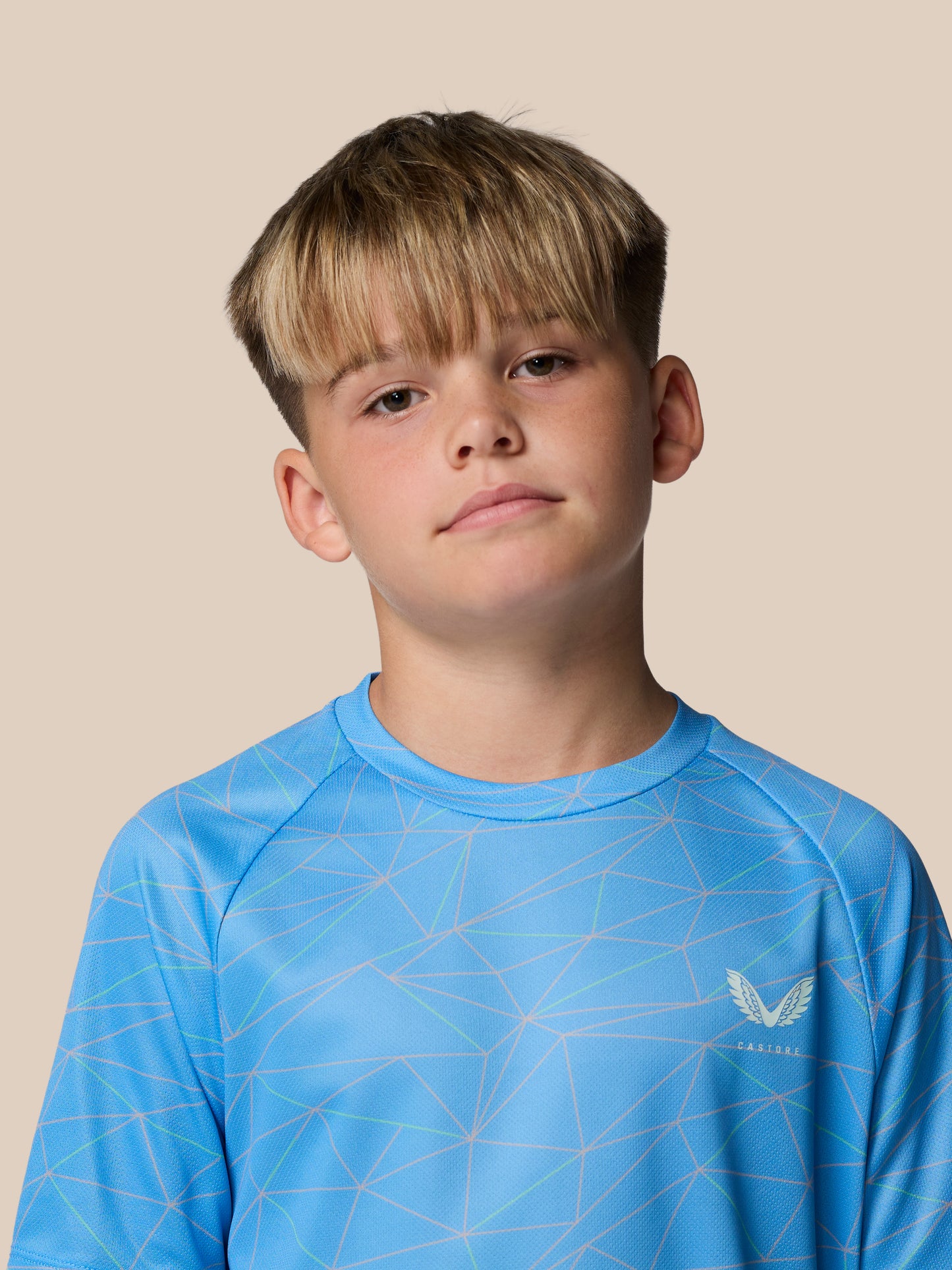 Young boy wearing a blue geometric print junior Castore T shirt against a beige studio backdrop.