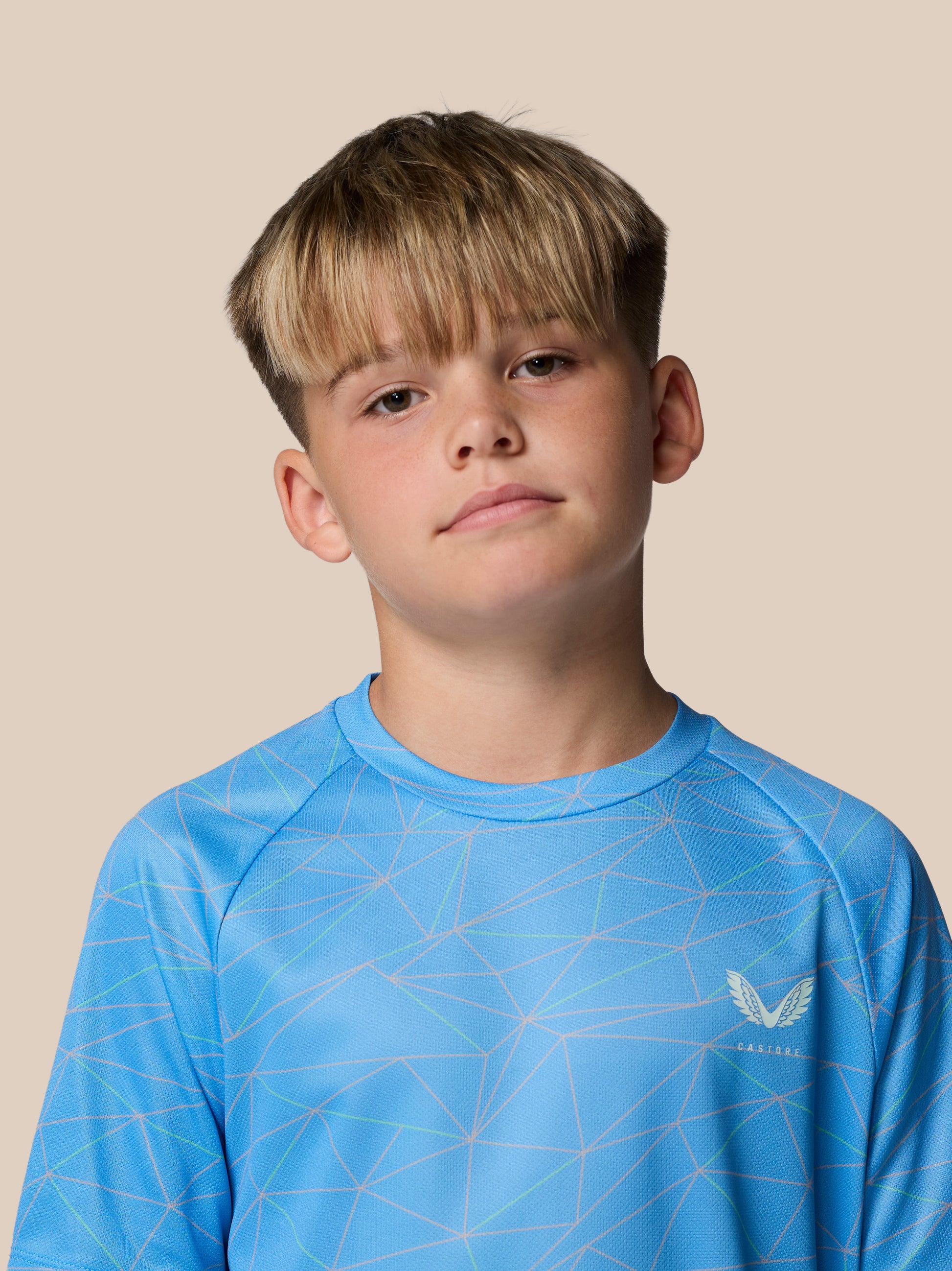 Young boy wearing a blue geometric print junior Castore T shirt against a beige studio backdrop.