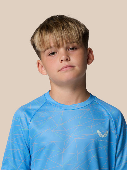 Young boy wearing a blue geometric print junior Castore T shirt against a beige studio backdrop.