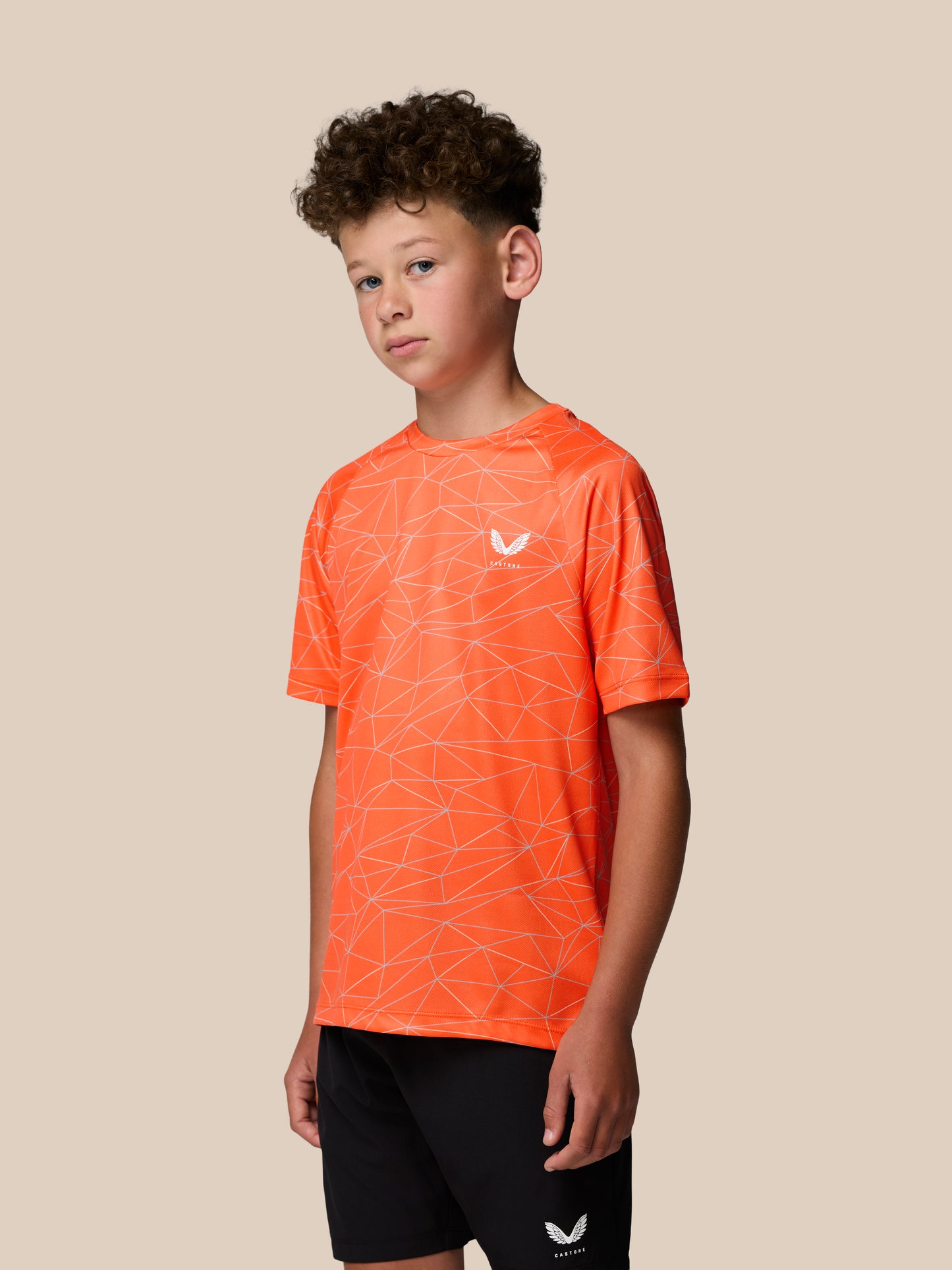 Young boy wearing bright orange short sleeve T shirt with white geometric line pattern and Castore logo on chest