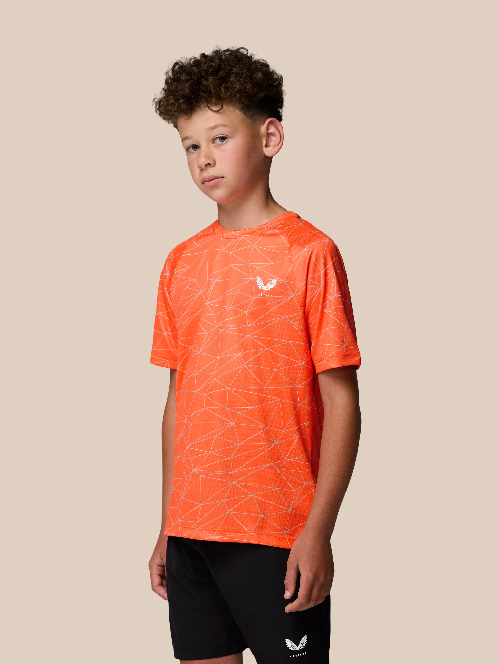 Young boy wearing bright orange short sleeve T shirt with white geometric line pattern and Castore logo on chest