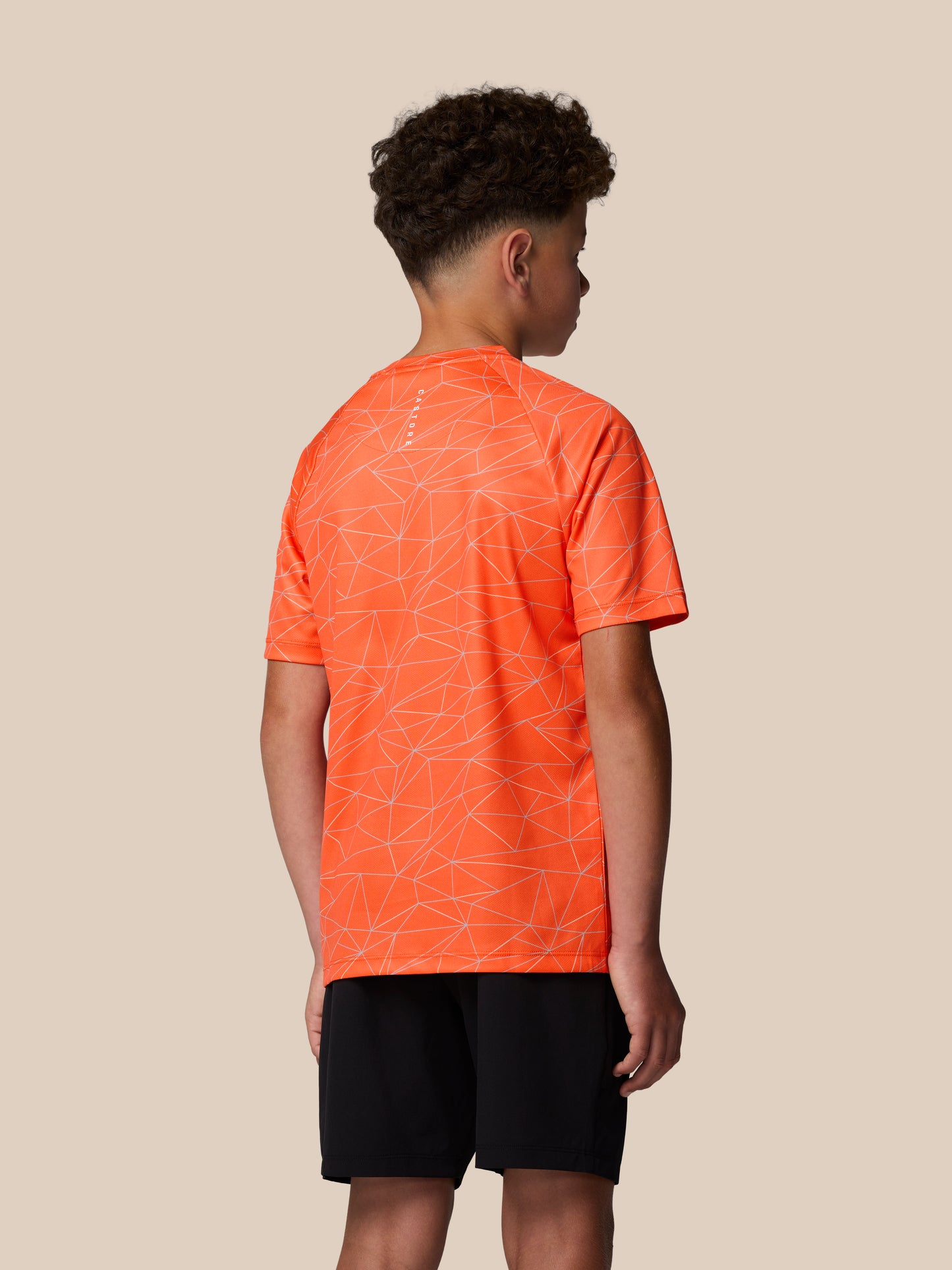 Back view of a young wearer in bright orange all over geometric print short sleeve T shirt with Castore logo on the back neck.