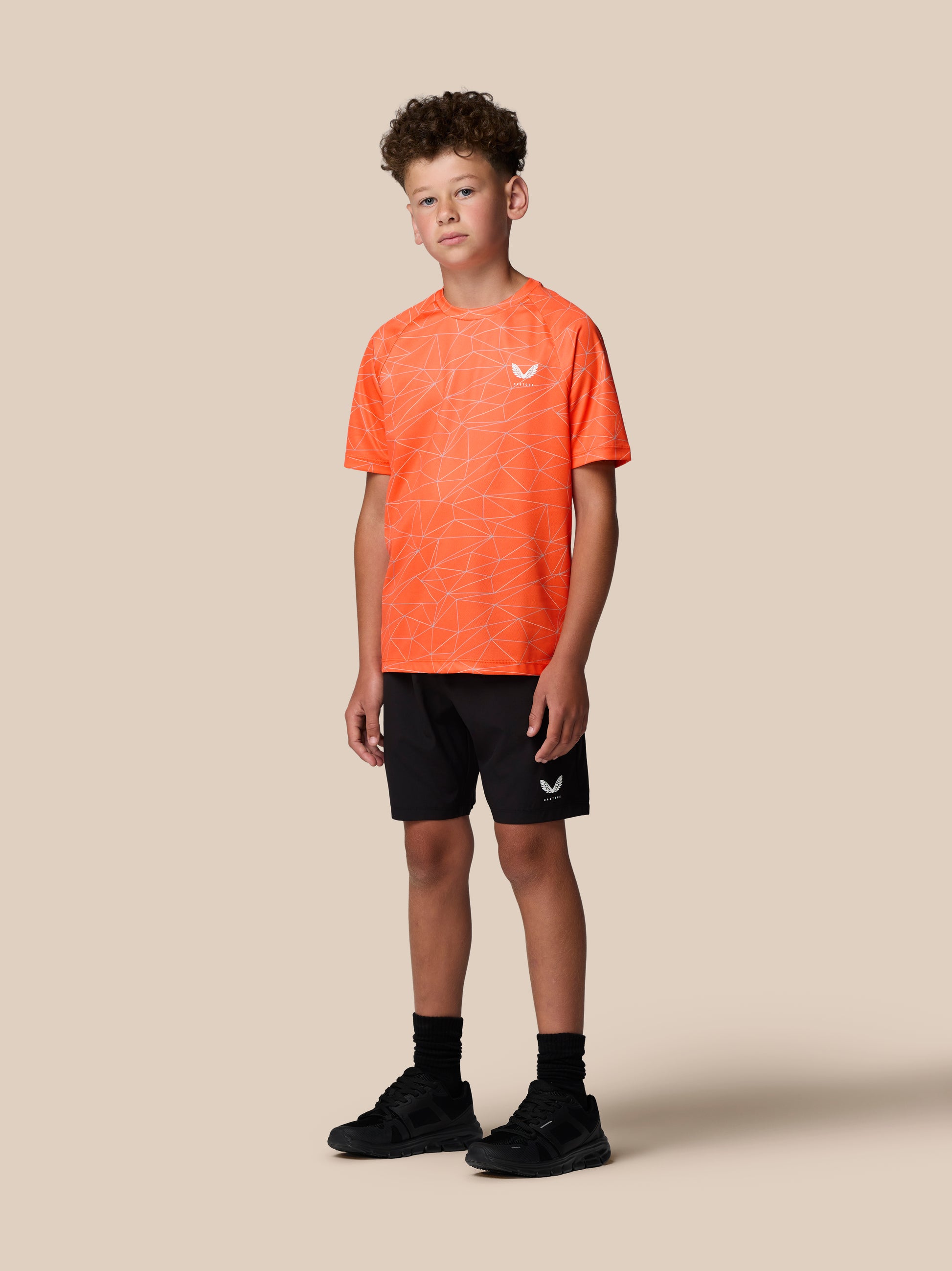 Young boy wearing bright orange allover print short sleeve T shirt with geometric white lines and Castore logo on left chest.