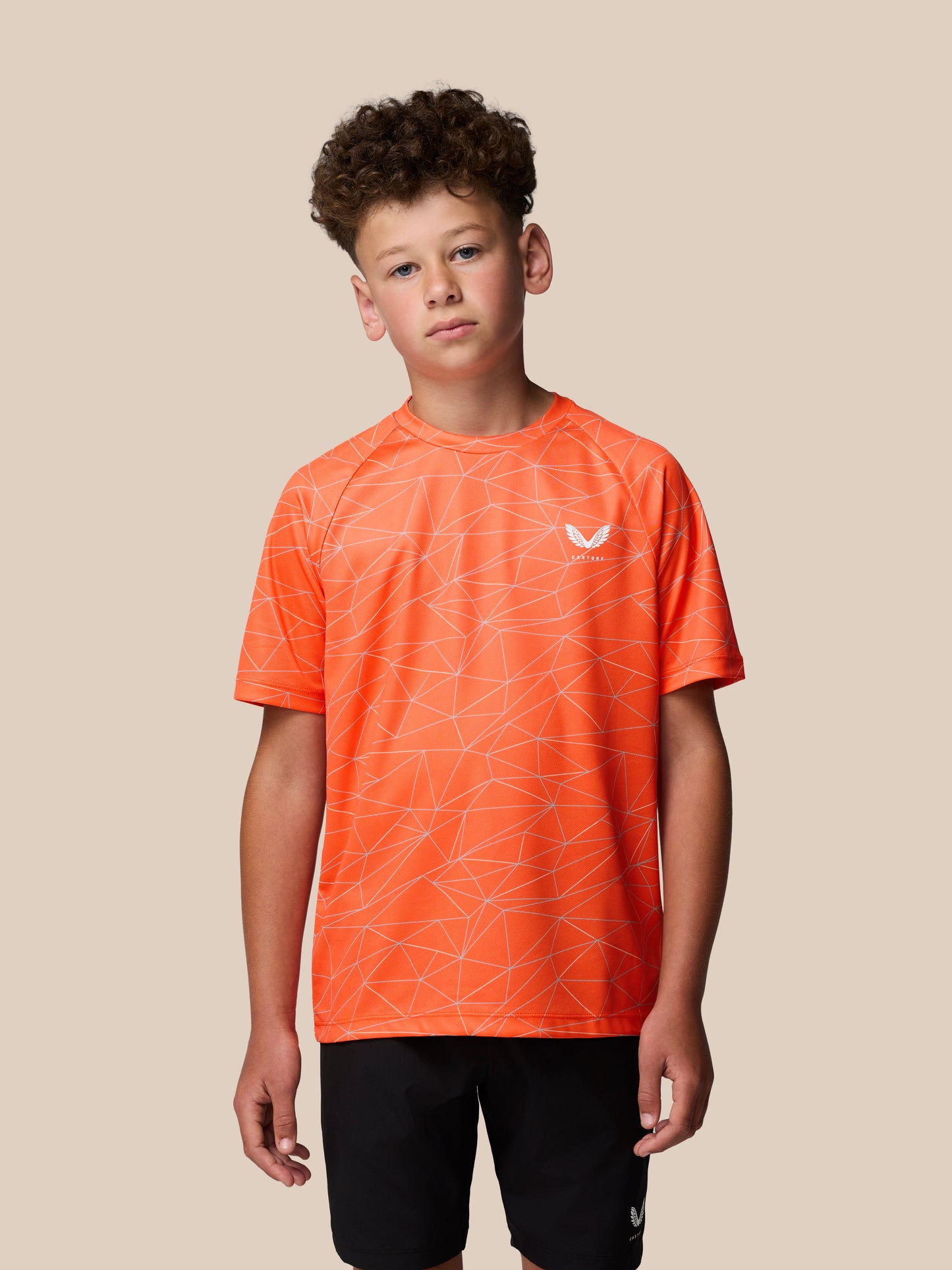 Young boy wearing bright orange short sleeve T shirt with white geometric all over print and Castore logo on the left chest