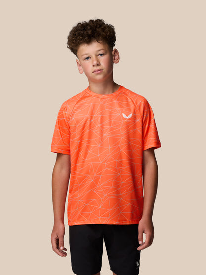 Young boy wearing bright orange short sleeve T shirt with white geometric all over print and Castore logo on the left chest