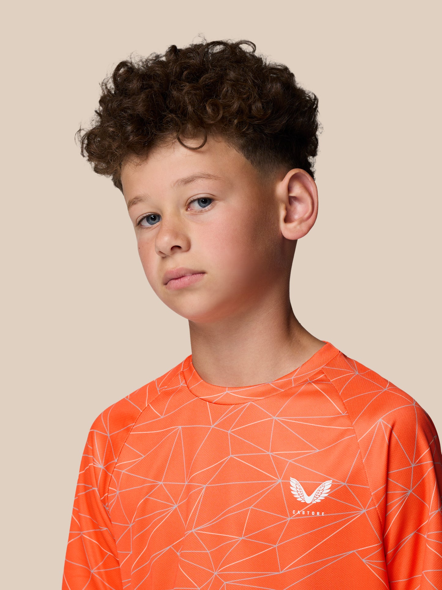 Portrait of a young boy wearing a bright orange geometric print short sleeve T shirt with Castore logo against a beige backdrop.
