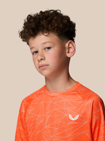 Portrait of a young boy wearing a bright orange geometric print short sleeve T shirt with Castore logo against a beige backdrop.