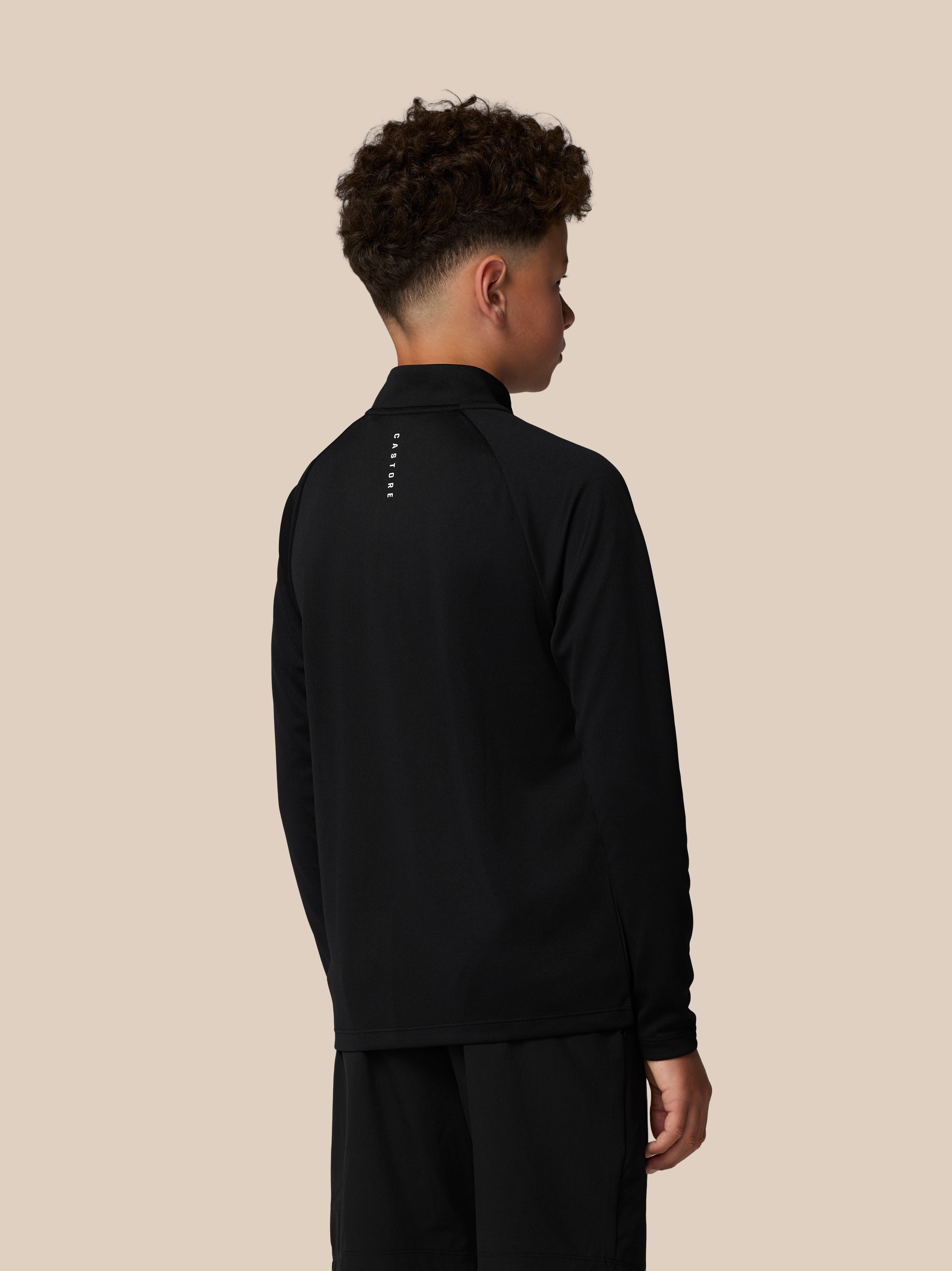 Three-quarter back view of a boy in a black quarter zip with branding along the neck and a small logo on the shorts.