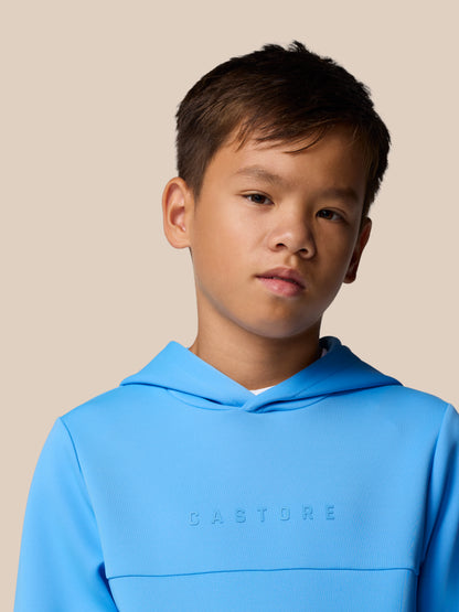Close-up of a young person wearing a bright blue Castore hoodie with an embossed chest logo against a beige background.
