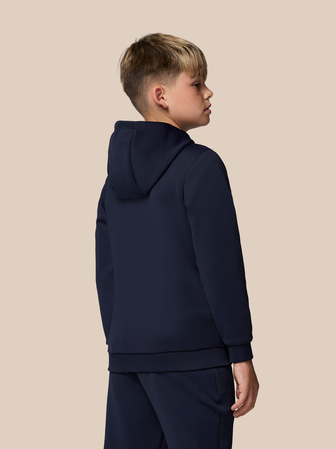 Boy seen from the back wearing navy Flex scuba jacket with hood against a beige backdrop