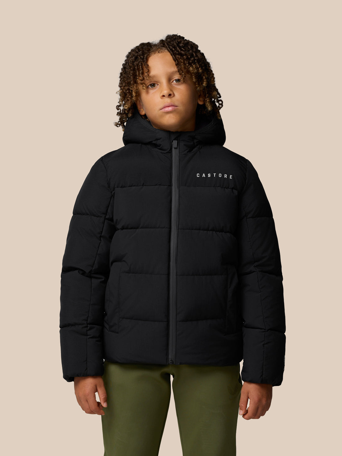 Child wearing black Quilts puffer jacket with hood and front zipper by Castore against a beige background