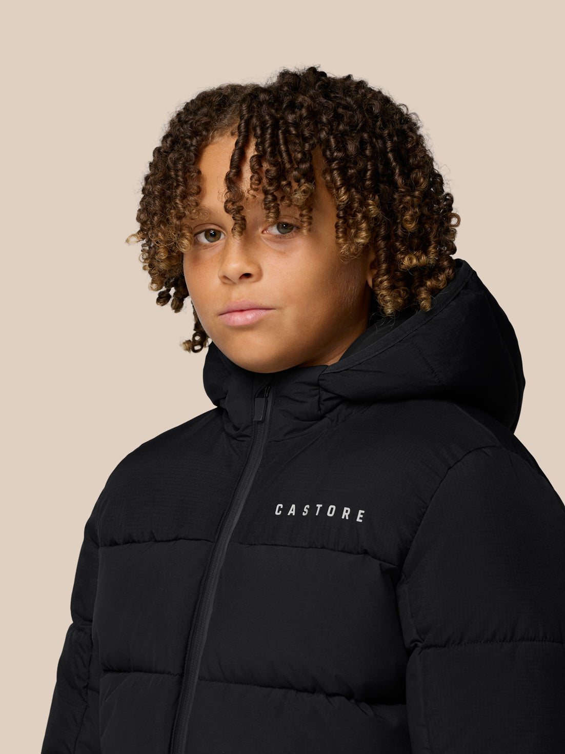 Close-up of a young boy wearing a black Castore Quilts puffer jacket with hood against a beige backdrop