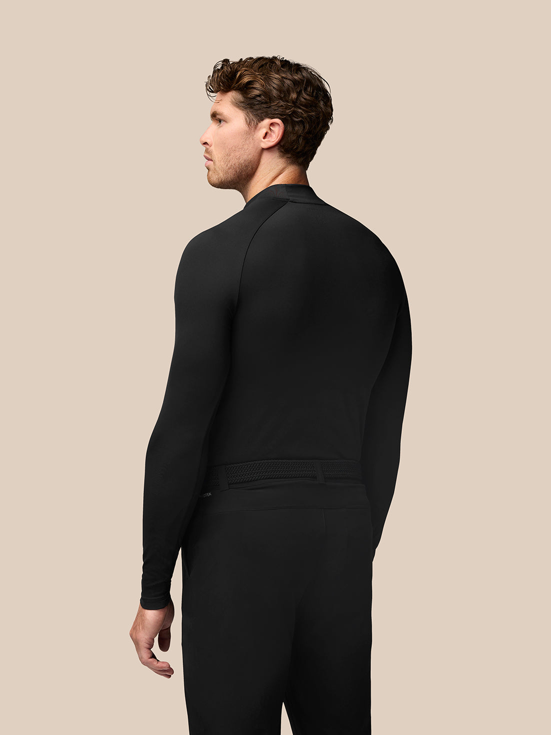 Back view of a man wearing a black golf long sleeve base layer with stand collar and raglan sleeves, Castore branding at the collar.