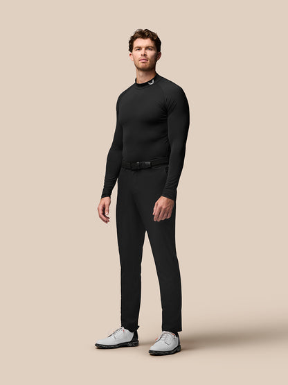 Full body shot of a man in a black golf long sleeve base layer with stand collar and raglan sleeves, belted trousers, Castore branding at the collar.