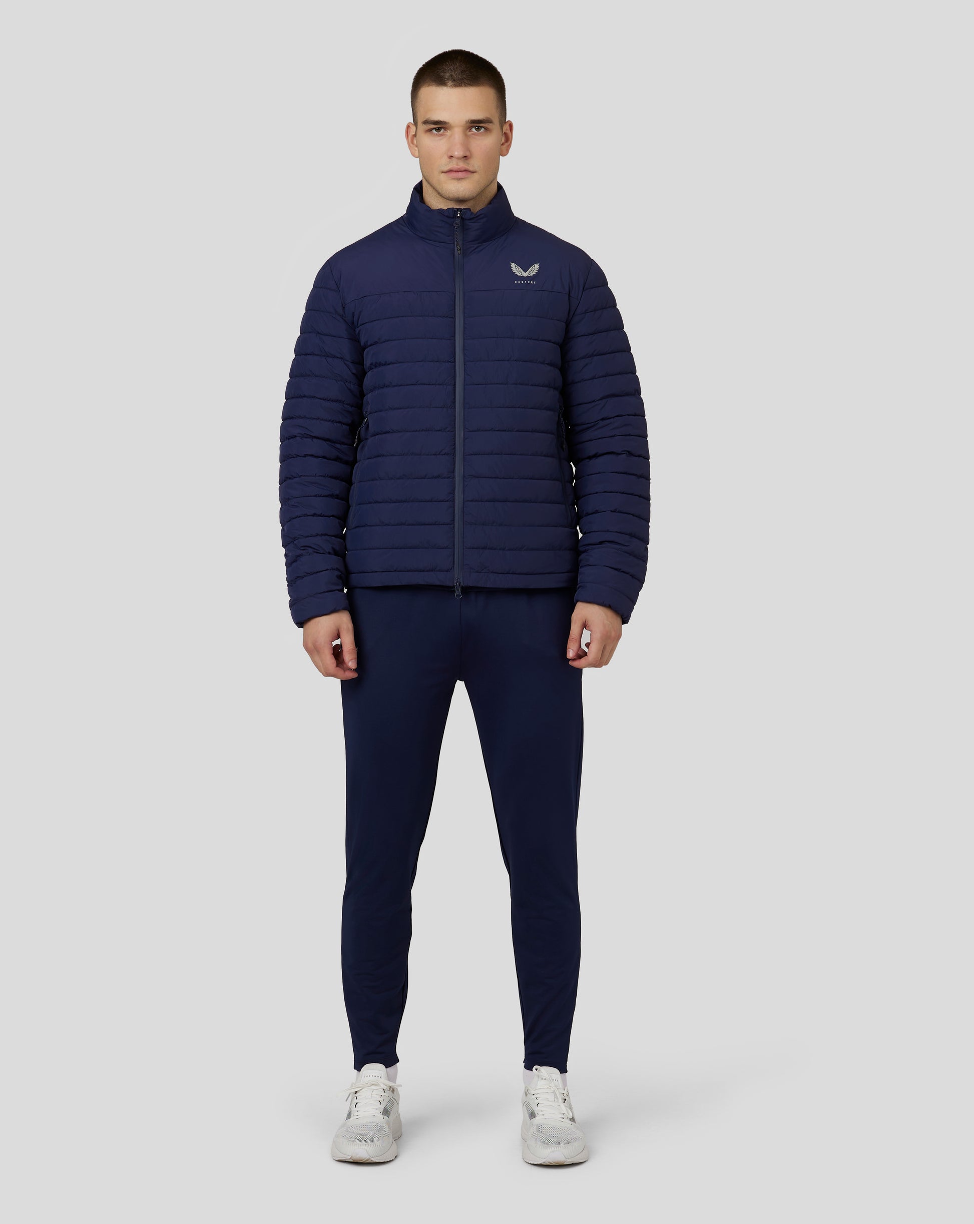 Male model wearing navy Castore puffer jacket with horizontal quilting, high stand collar, front zip and left chest logo