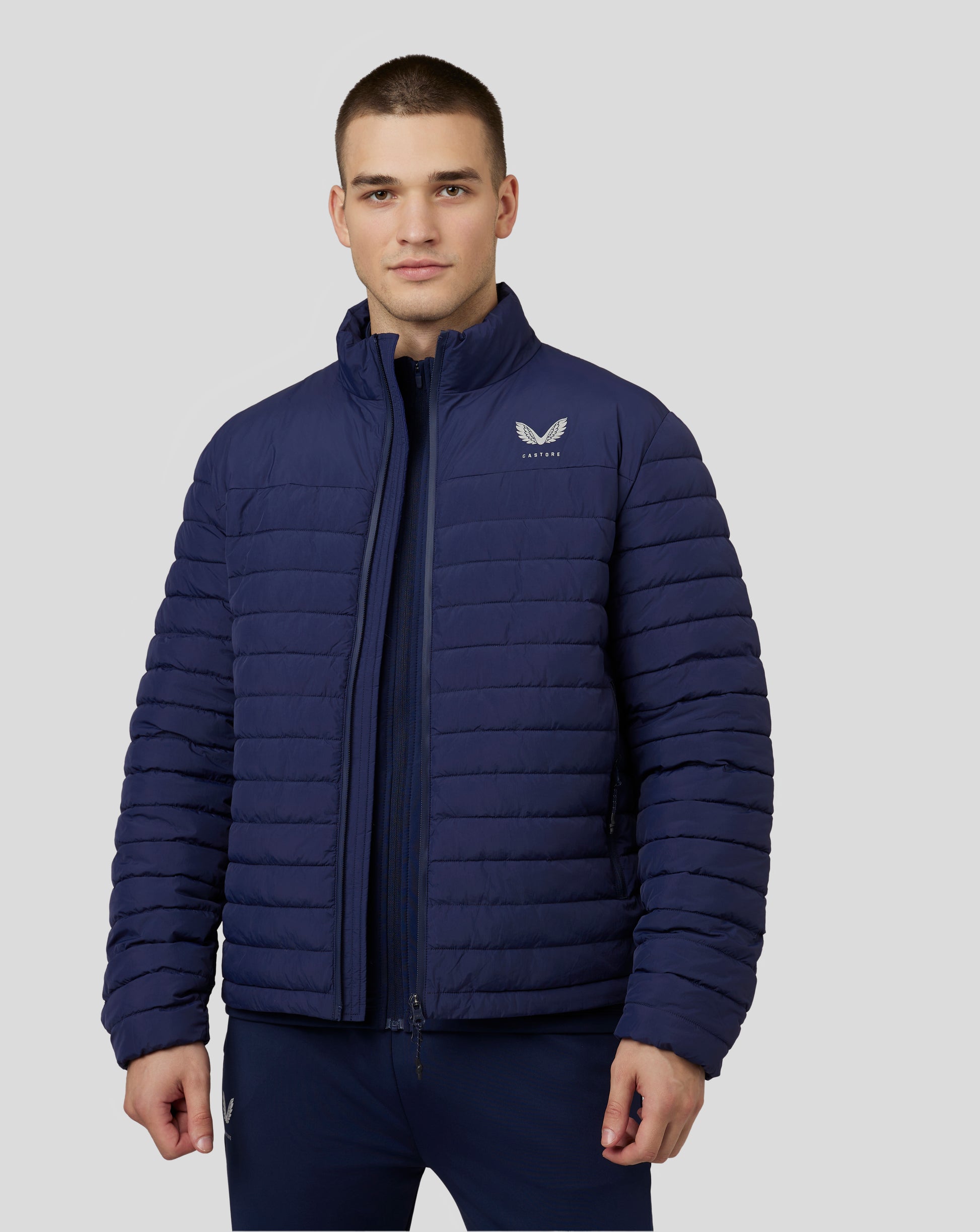 Male model wearing navy quilted puffer jacket with high stand collar and zip front, Castore logo on left chest