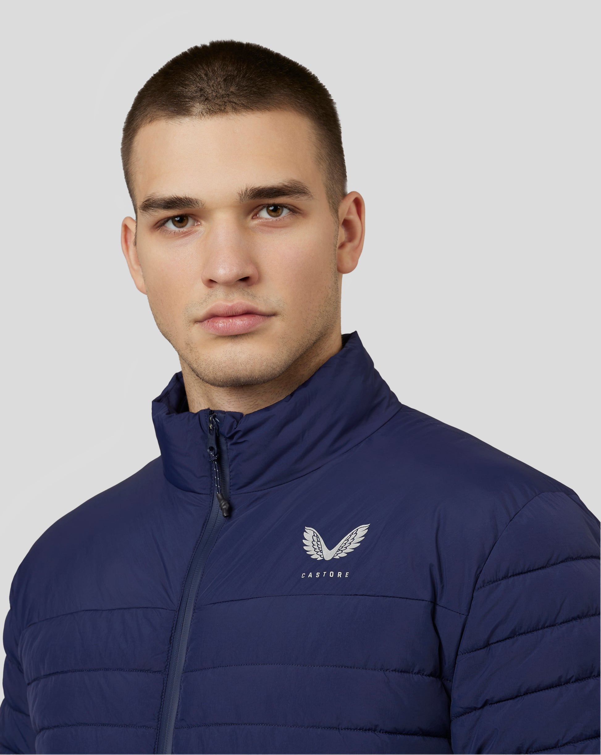 Close-up of a man wearing a navy Castore travel puffer jacket with front zipper and wing emblem