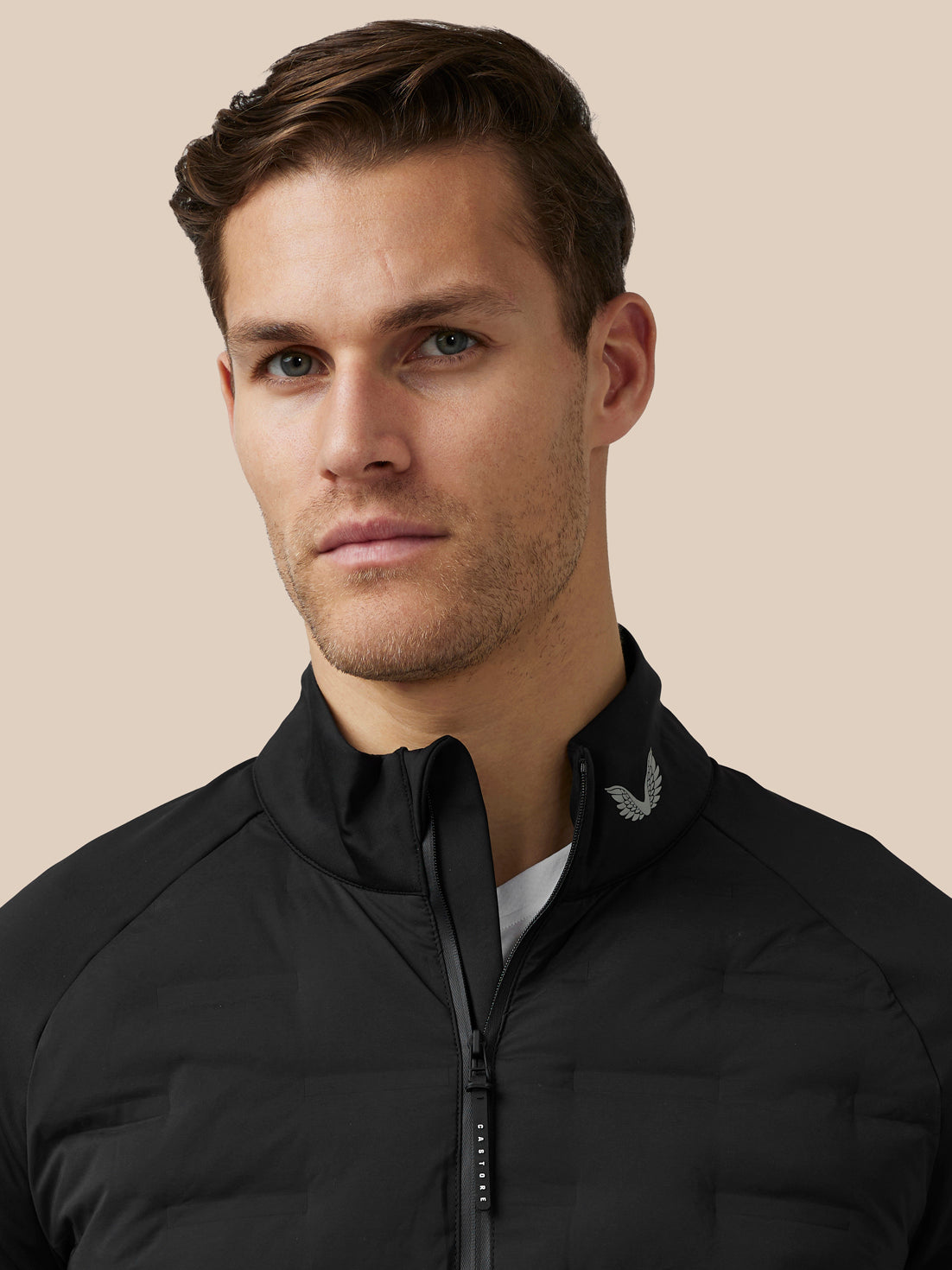 Close-up of a male model in a black zip jacket with stand collar and a small wing logo on the collar.