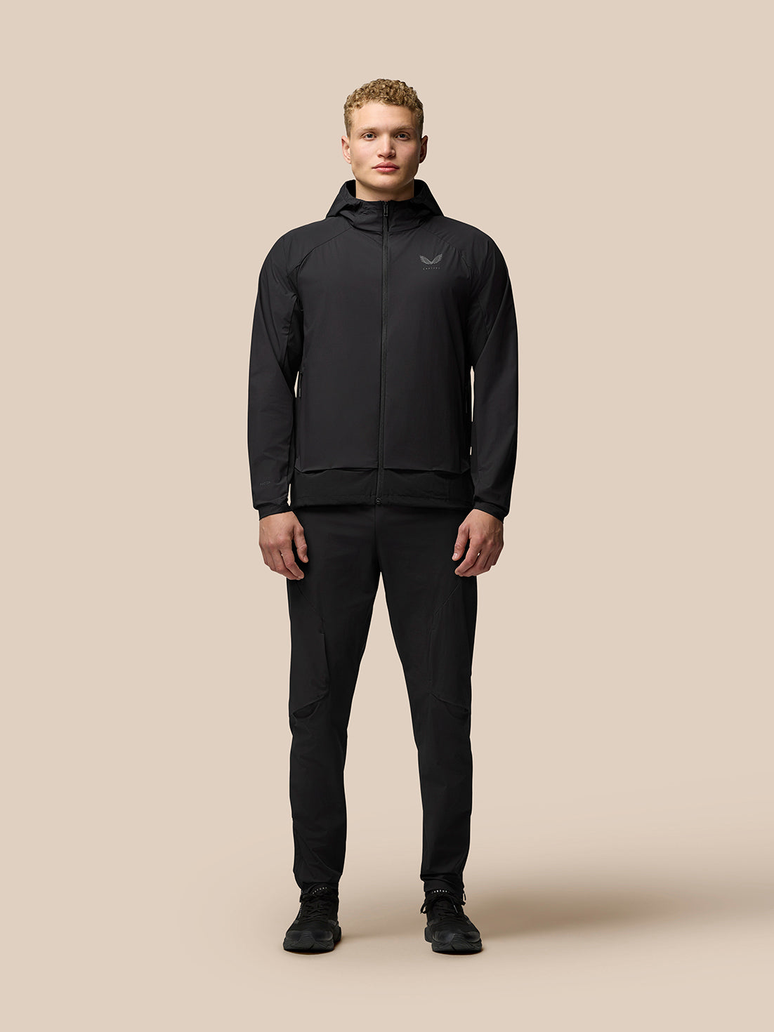 Full body shot of a man in black Flex Woven Joggers and jacket against a beige background