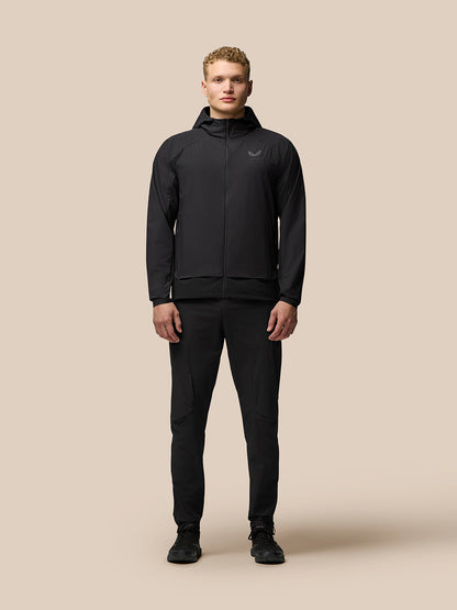 Full body shot of a man in black Flex Woven Joggers and jacket against a beige background