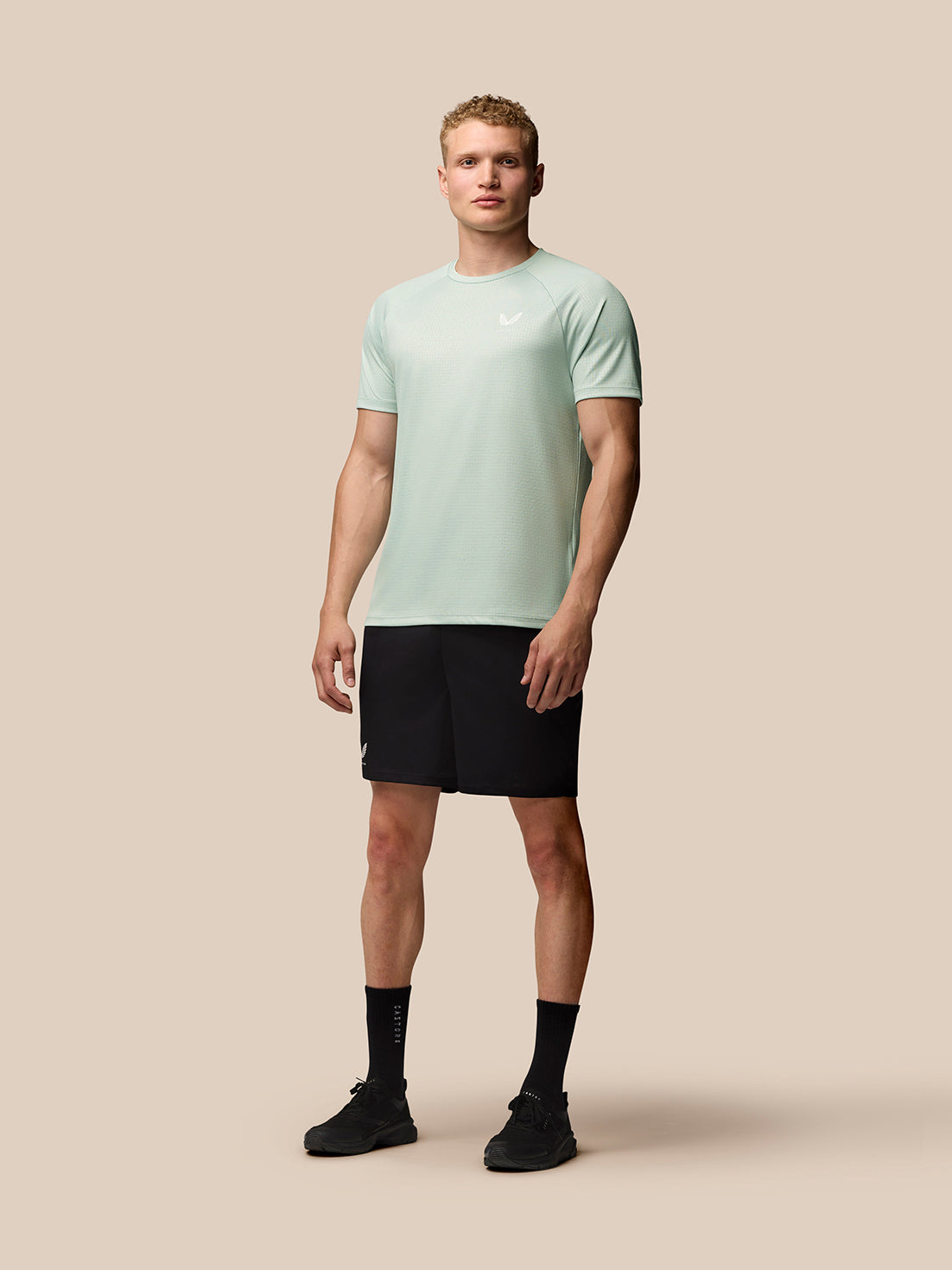 Model wearing mint green Adapt Training T Shirt with black shorts, standing against a beige background
