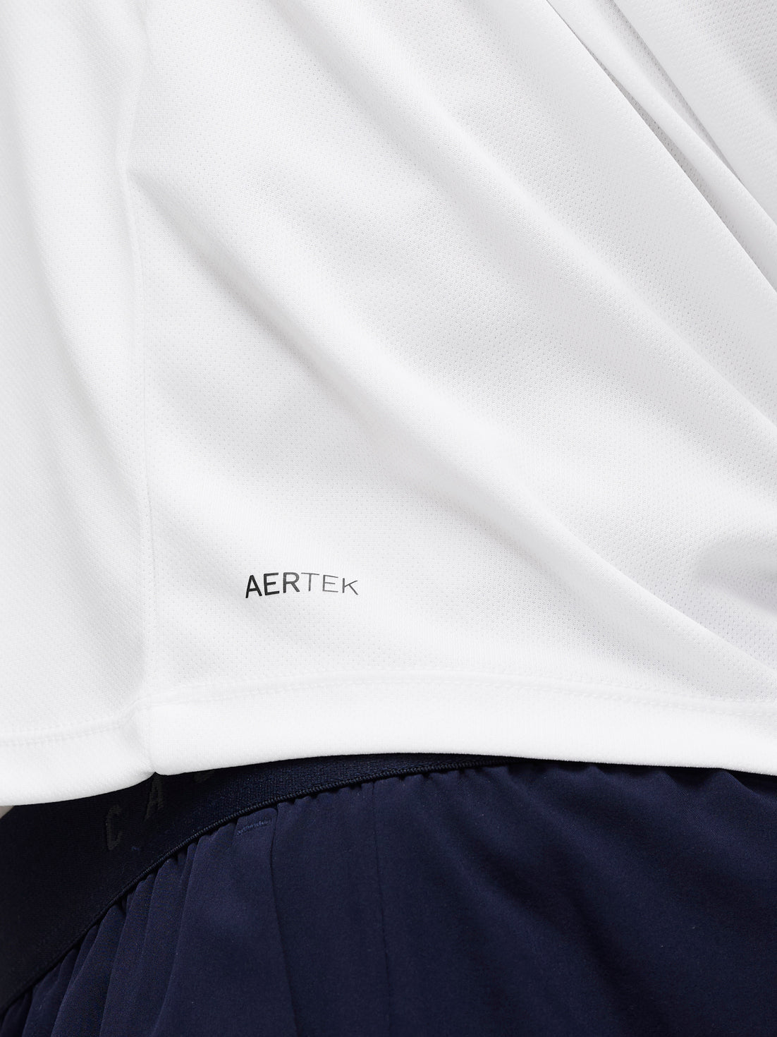 Close up of white Adapt Training T Shirt fabric with AERTEK branding near the hem, navy shorts visible in the background