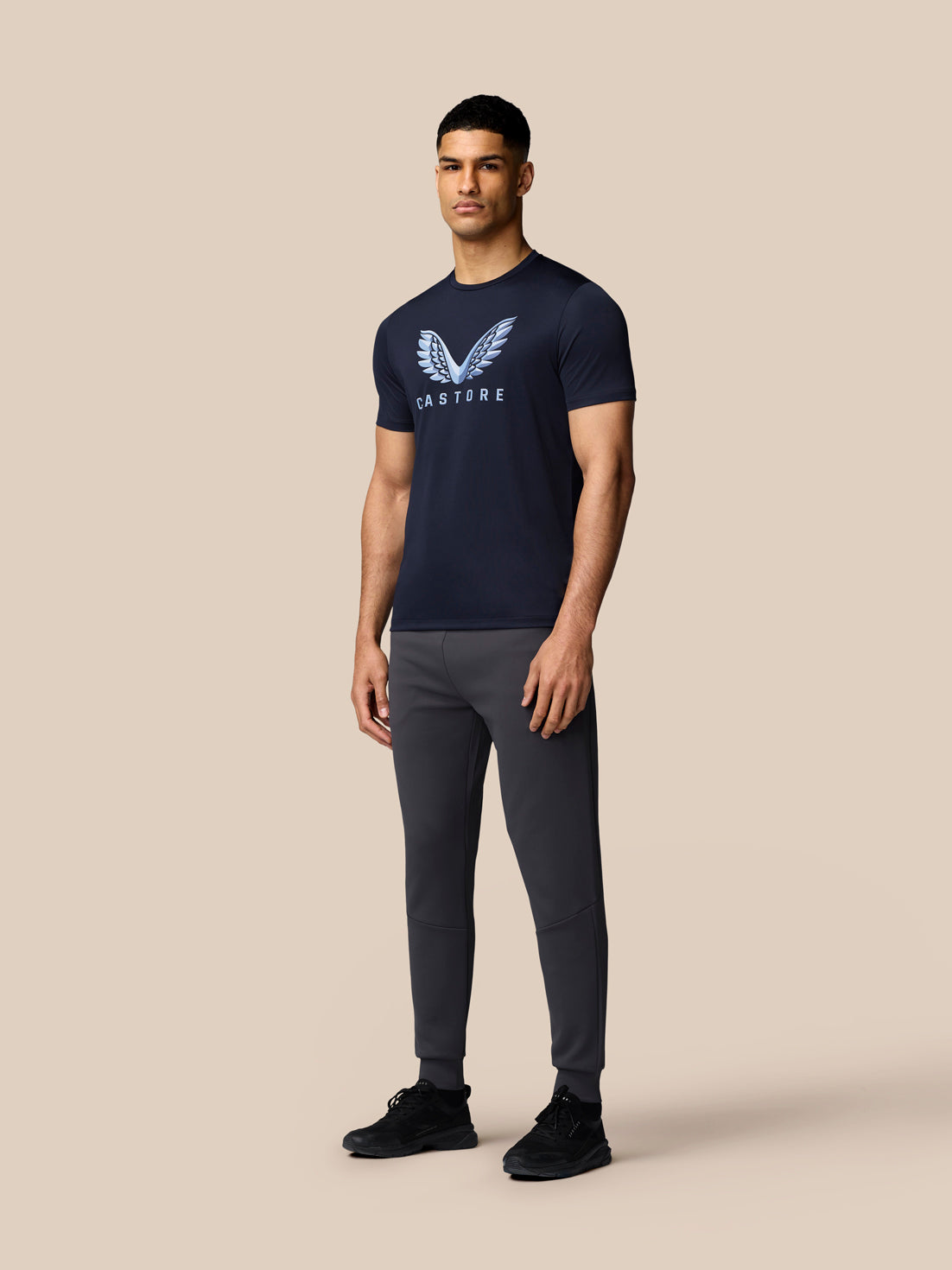 Male model wearing navy graphic T-shirt with Castore wings logo on chest and gray joggers